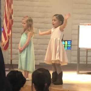 Two little girls, one in a light blue dress and one in a pink dress. The girl in pink is gesturing broadly while an audience looks on.