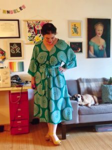 A woman wears a teal-colored midi-length dress while a dog watches from the couch.