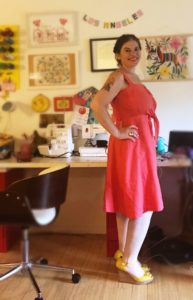 A woman stand in profile, smiling at the camera while wearing a coral-colored sundress.