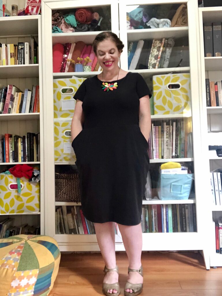 A white woman with dark hair wears a black dress and a colorful necklace in front of a colorful bookshelf.