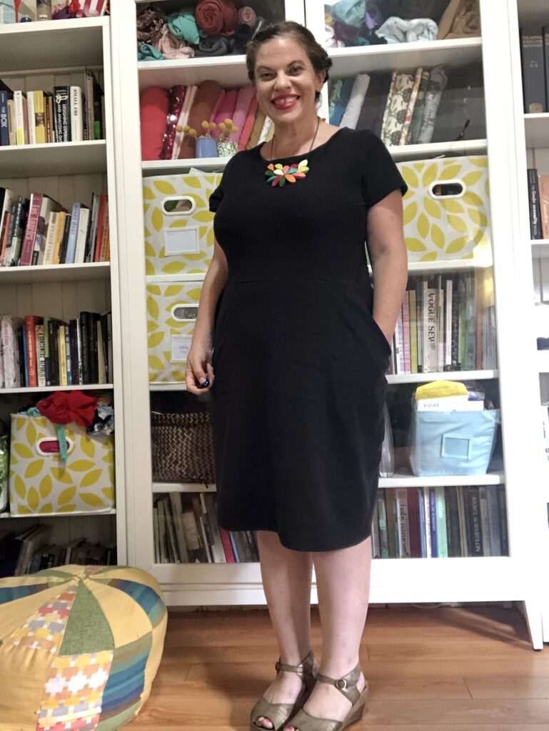 A white woman with dark hair wears a black dress and a colorful necklace while standing in front of a colorful bookshelf.