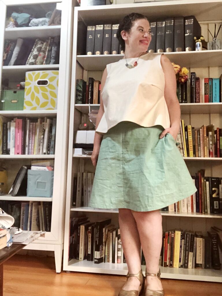 A woman wears a light-blue skirt and a cream-colored tank in front of a colorful bookshelf.