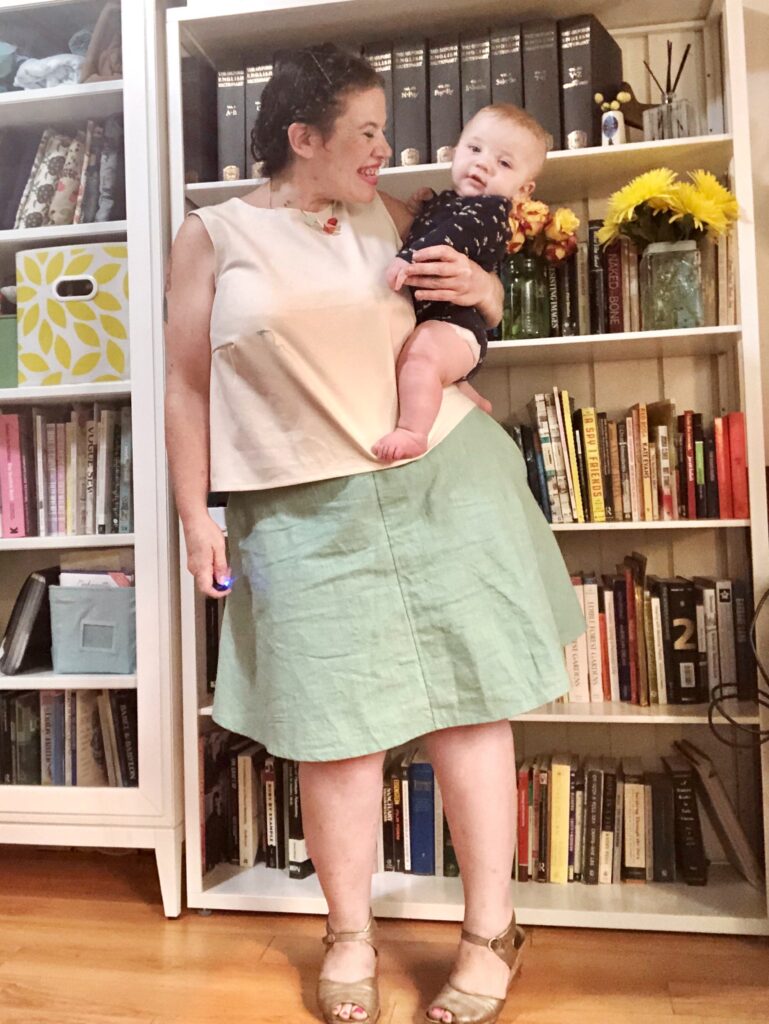 A woman holds a fat baby while wearing a blue skirt and white top in front of a colorful bookshelf.