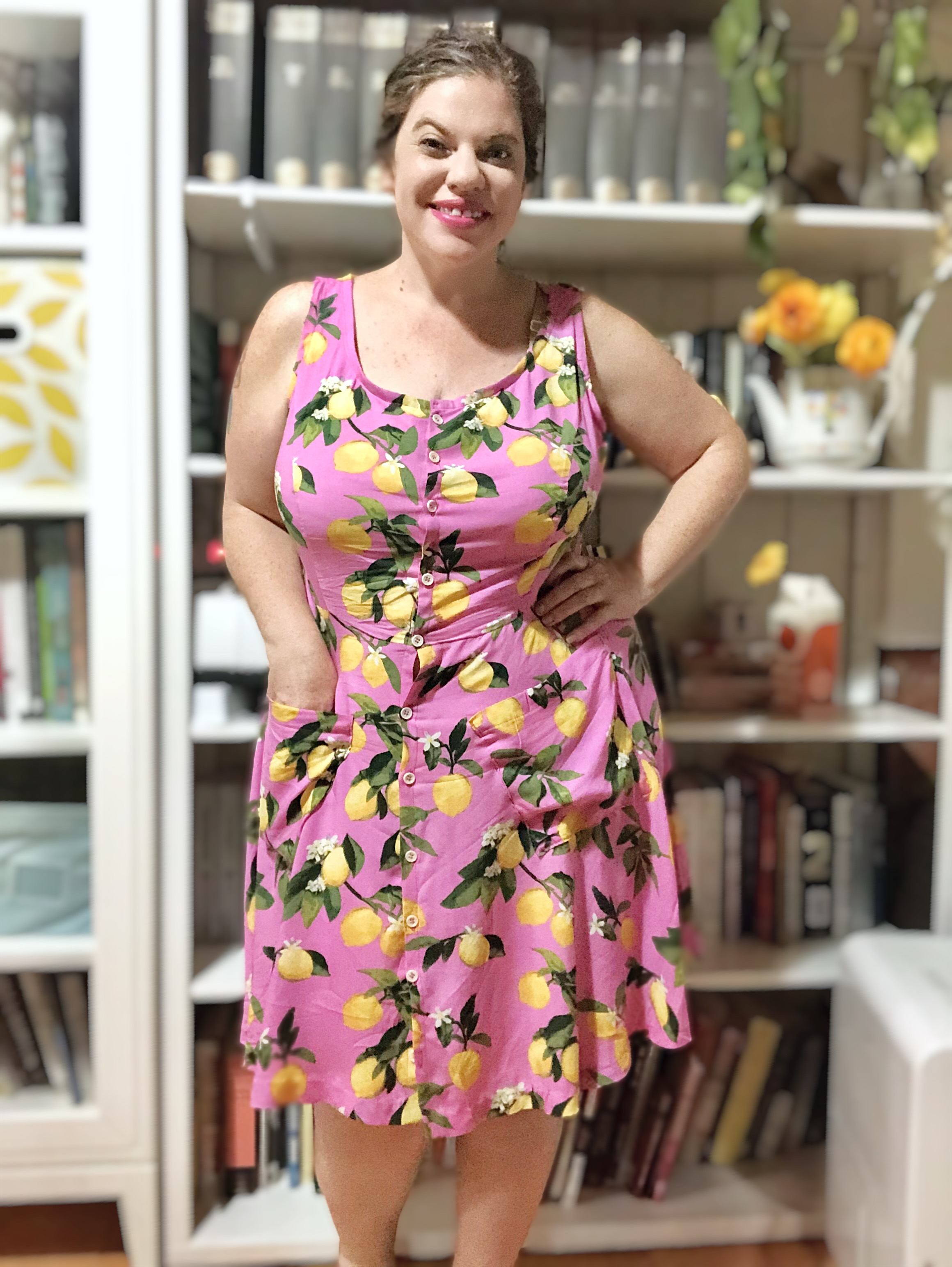 White woman with dark hair stands in front of a colorful bookshelf wearing a pink sundress decorated with yellow lemons.