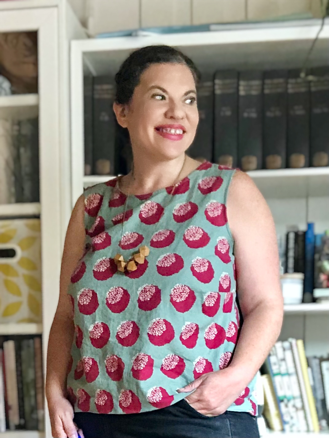 White woman with dark hair stands in front of a bookshelf wearing a blue tank top with large red dots, as well as a yellow wooden necklace.