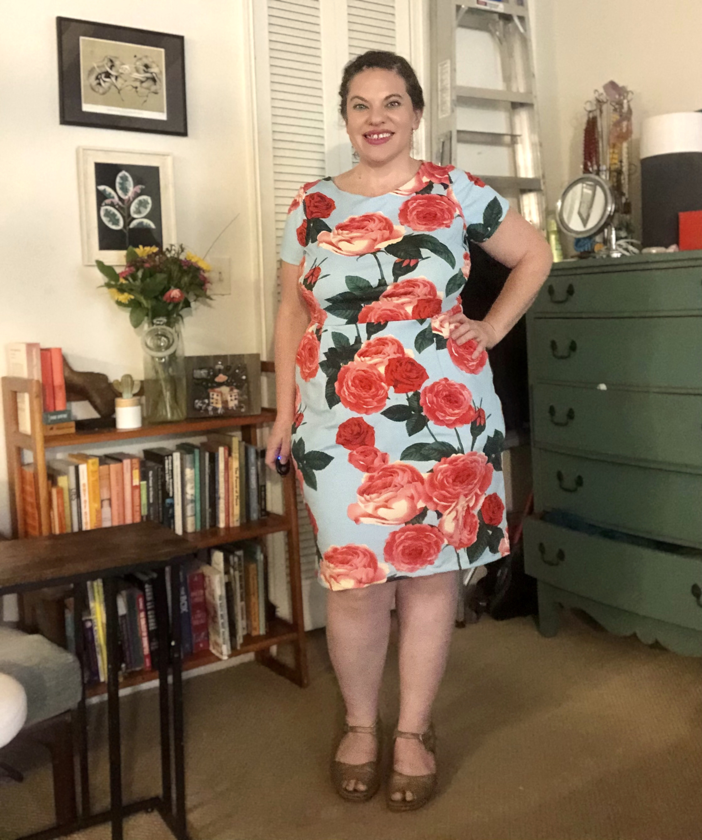 A woman stands in front of a bookshelf wearing a blue dress with red roses scattered across.