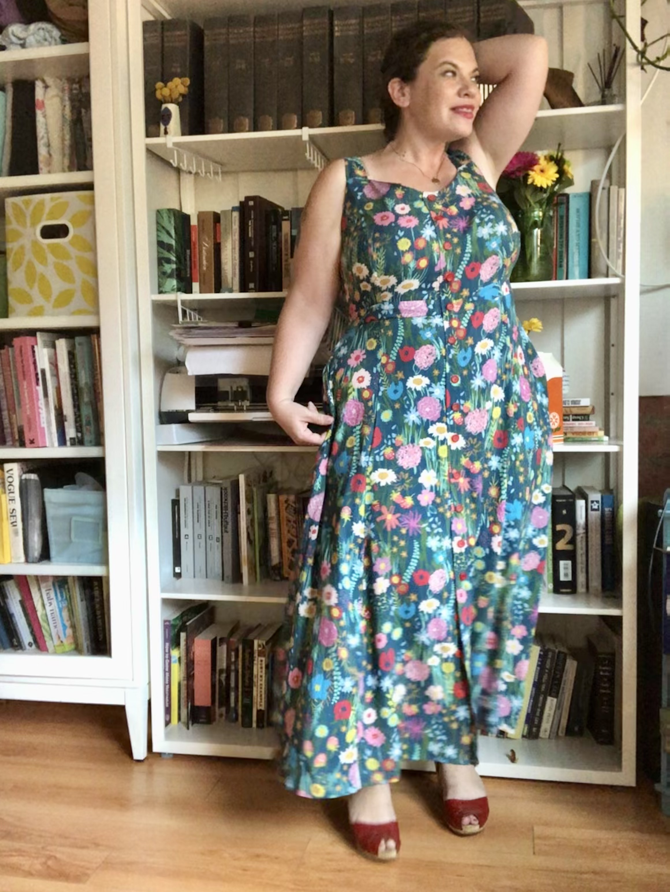 A white woman with dark hair stands in front of a bookshelf wearing a colorful floral maxi dress.
