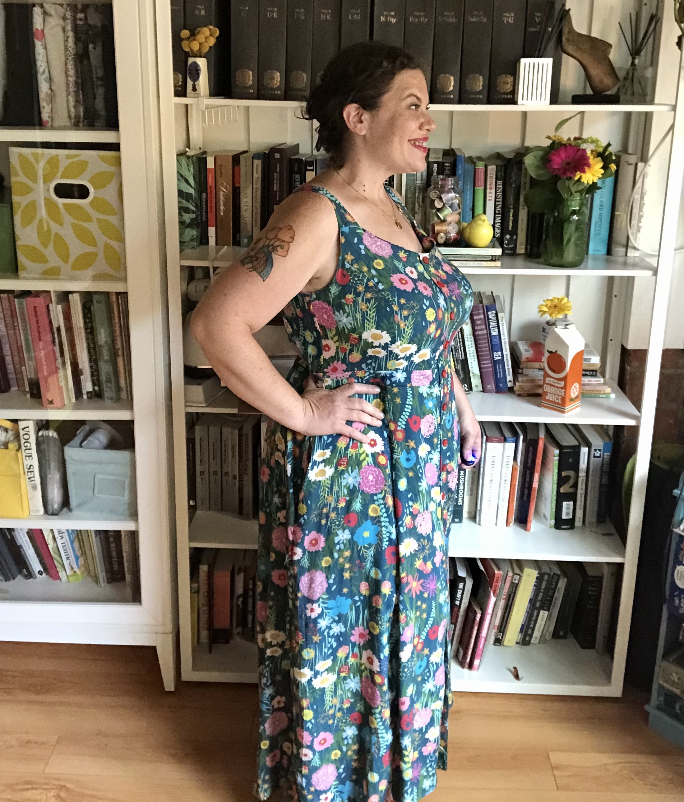 A white woman with dark hair stands in profile in front of a bookshelf, wearing a colorful floral maxi dress. A tattoo is slightly visible on the upper part of her right arm.