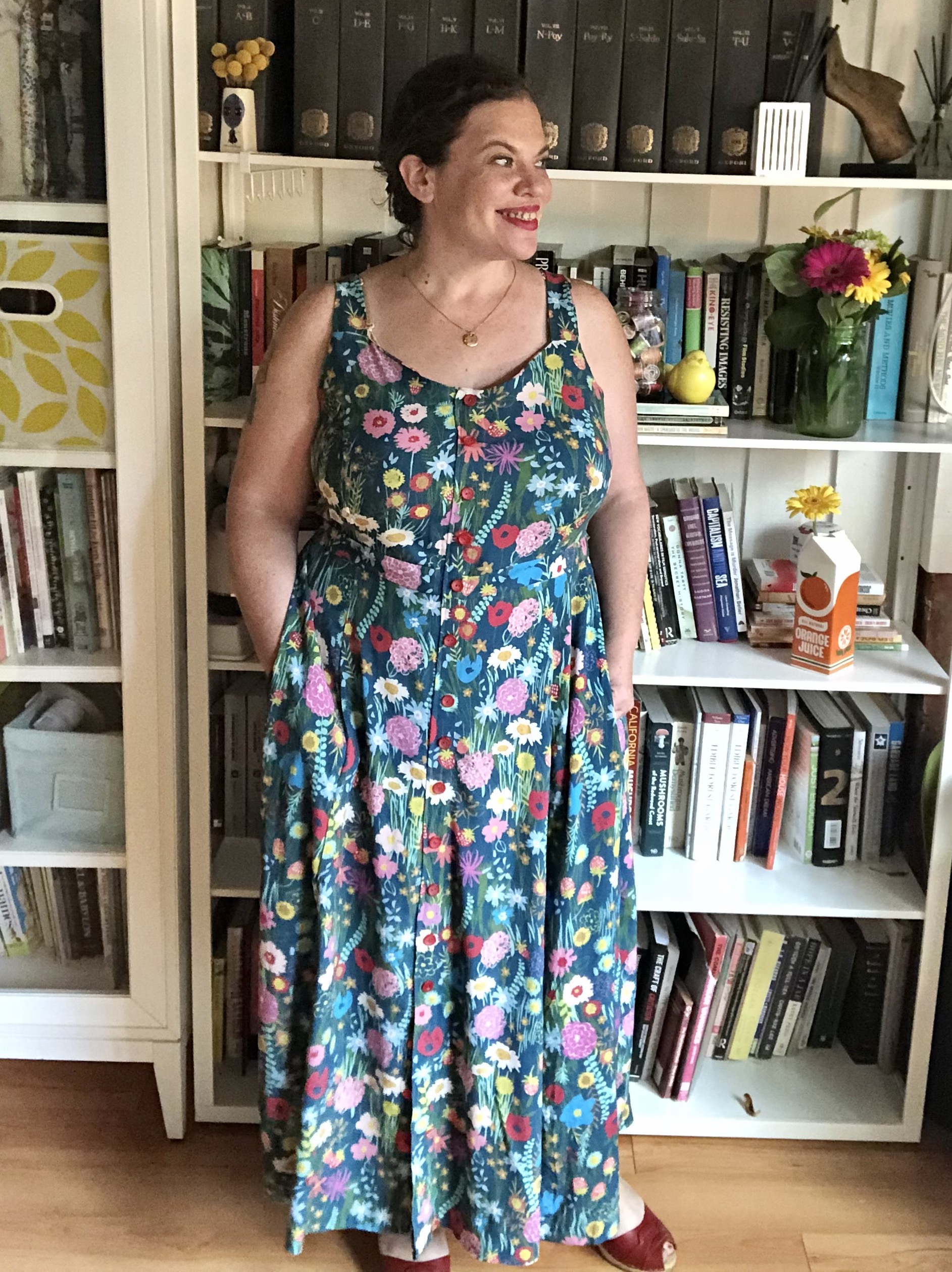 A white woman with dark hair stands in front of a bookshelf wearing a flowered maxi dress.