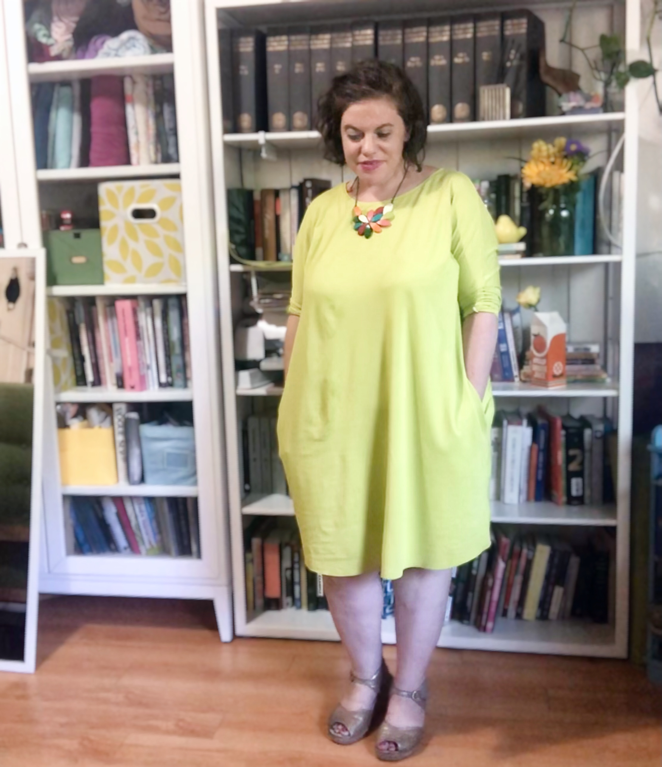 A white woman with dark hair stands in front of a bookshelf, wearing a bright green, baggy dress, her hands in her pockets.